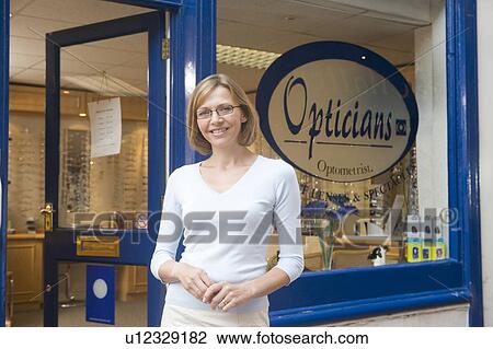 Stock Image - Woman standing at front entrance of optometrists smiling. Fotosearch