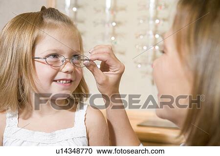 Stock Image - Woman trying eyeglasses on young girl at optometrists smiling. Fotosearch