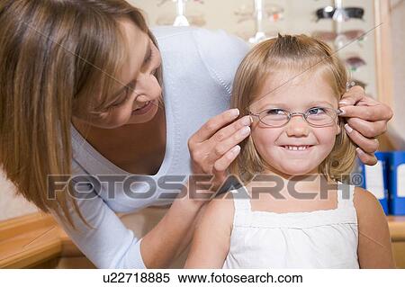 Stock Photography - Woman trying eyeglasses on young girl at optometrists smiling. Fotosearch