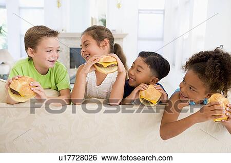 Stock Photograph - Four young children eating cheeseburgers in living room smiling. Fotosearch