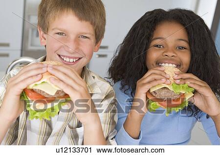 Stock Image - Two young children in kitchen eating cheeseburgers smiling. Fotosearch