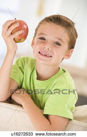 Stock Image - Young boy eating apple in living room smiling. Fotosearch