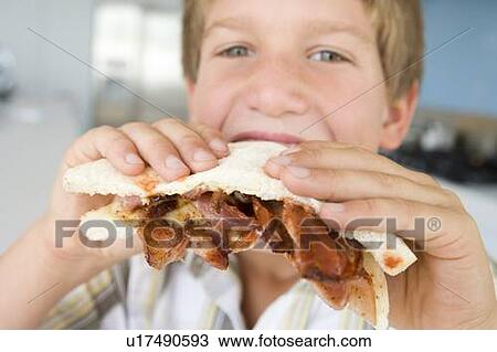 Stock Image - Young boy in kitchen eating bacon sandwich. Fotosearch