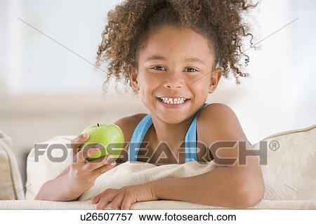 Young girl eating apple in living room smiling View Large Photo Image Stock Photo - Young girl eating apple in living room smiling. Fotosearch