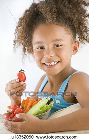 Stock Image - Young girl eating bowl of vegetables in living room smiling. Fotosearch