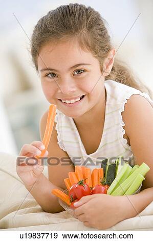 Stock Photo - Young girl eating bowl of vegetables in living room smiling. Fotosearch