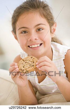 Stock Image - Young girl eating cookie in living room smiling. Fotosearch