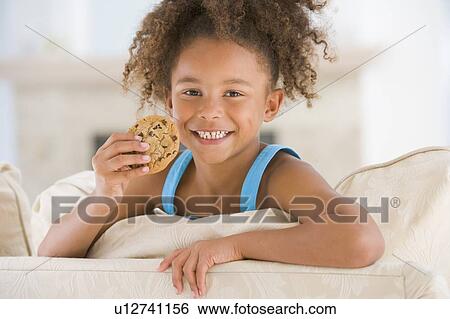 Stock Photograph - Young girl eating cookie in living room smiling. Fotosearch