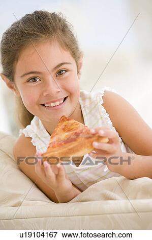 Stock Photo - Young girl eating pizza slice in living room smiling. Fotosearch