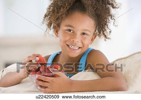 Stock Photo - Young girl eating strawberries in living room smiling. Fotosearch