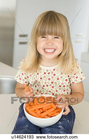 Stock Image - Young girl in kitchen eating carrot sticks smiling. Fotosearch