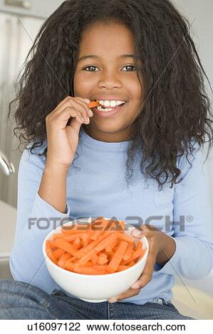Stock Image - Young girl in kitchen eating carrot sticks smiling. Fotosearch