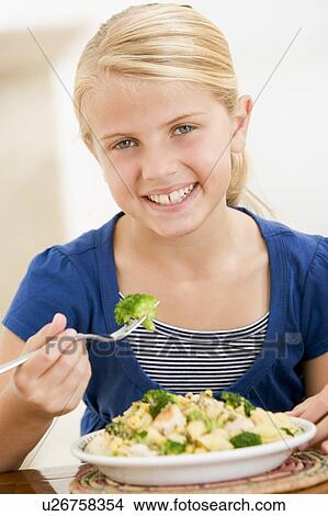 Picture - Young girl indoors eating pasta with brocolli smiling. Fotosearch