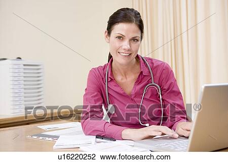 Stock Image - Doctor using laptop in doctor's office smiling. Fotosearch