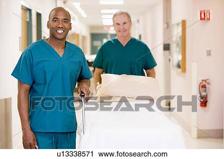 Stock Image - Two Orderlies Pushing An Empty Bed Down A Hospital Corridor. Fotosearch