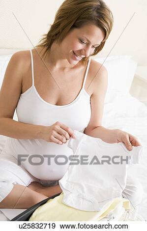 Stock Photo - Pregnant woman packing baby clothing in suitcase smiling. Fotosearch