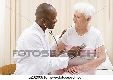 Stock Photograph - Doctor giving checkup with stethoscope to woman in exam room smiling. Fotosearch