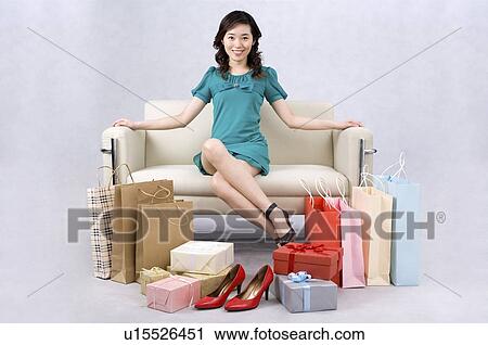 Stock Image - Young woman sitting on sofa surrounded by shopping gains. Fotosearch