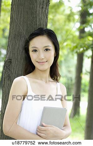 Young woman holding book leaning against tree View Large Photo Image Stock Image - Young woman holding book leaning against tree. Fotosearch
