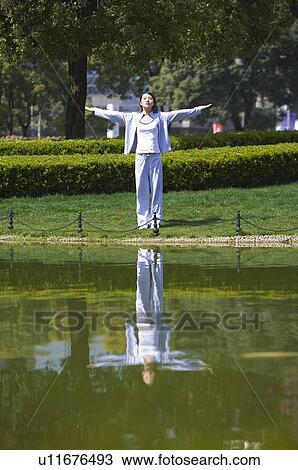 Stock Image - Young woman with arms outstretched standing at water's edge. Fotosearch