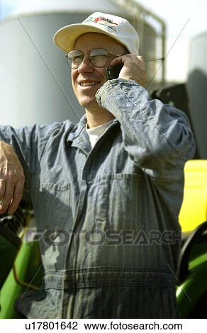 Stock Image - Man leaning against tractor talking on cell phone. Fotosearch