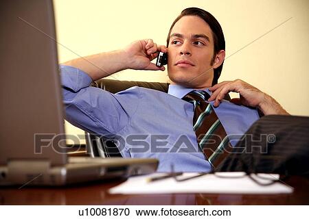 Young businessman sitting at desk and talking on cell phone View Large Photo Image Stock Image - Young businessman sitting at desk and talking on cell phone. Fotosearch