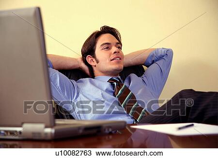Stock Image - Young businessman sitting at desk with arms raised. Fotosearch