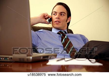 Stock Photography - Young businessman sitting at desk and talking on cell phone. Fotosearch