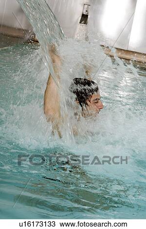 Stock Image - Young man standing under water spray in spa pool. Fotosearch