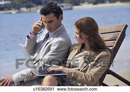 Stock Image - Business couple on beach with cell phone and laptop. Fotosearch