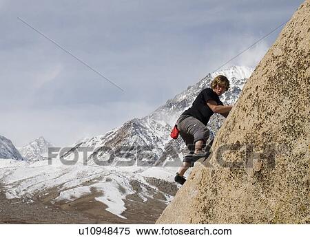 Stock Photography - Man (20-25) climbing a rock face with a snow-capped mountain in the background, Bishop, California, USA. Fotosearch