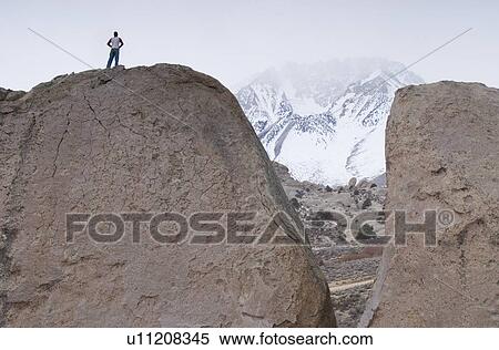 Stock Photography - Man (20-25) standing on top of a large boulder with a snow-capped mountain in the distance partially hidden by fog. Bishop, California. Fotosearch