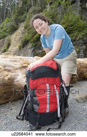 Stock Image - Woman (20-25) packing a large backpack on a beach with a large beach log in the background, West Coast Trail, Pacific Rim National Park Reserve, BC. Fotosearch