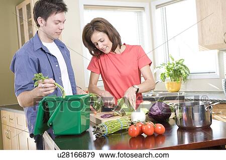 Stock Photo - Man places carrot top into kitchen compost bin as woman cuts vegetables, Winnipeg, Canada. Fotosearch