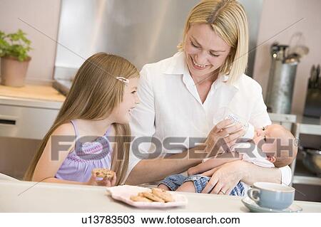 Stock Image - Mother feeding baby in kitchen with daughter eating cookies and smiling. Fotosearch