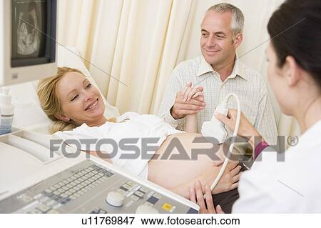 Stock Photo - Pregnant woman getting ultrasound from doctor with husband watching. Fotosearch