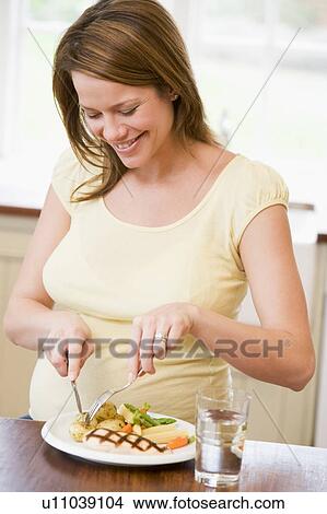 Picture - Pregnant woman in kitchen eating chicken and vegetables smiling. Fotosearch