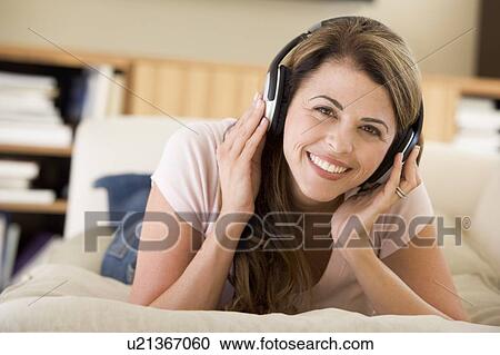 Stock Image - Woman in living room listening to headphones smiling. Fotosearch