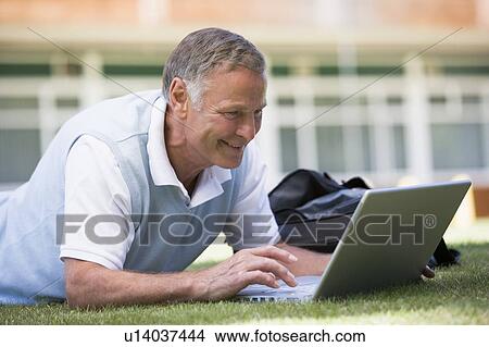 Picture - Man lying on lawn of school with laptop. Fotosearch