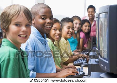 Stock Image - Six children at computer terminals with teacher in background (selective focus/high key). Fotosearch