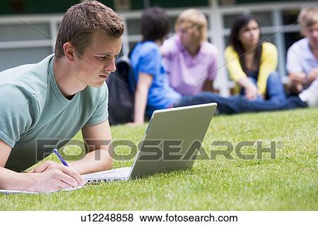 Stock Photo - Student outdoors on lawn using laptop with other students in background (selective focus). Fotosearch