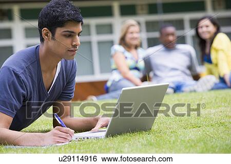 Stock Photograph - Student outdoors on lawn using laptop with other students in background (selective focus). Fotosearch