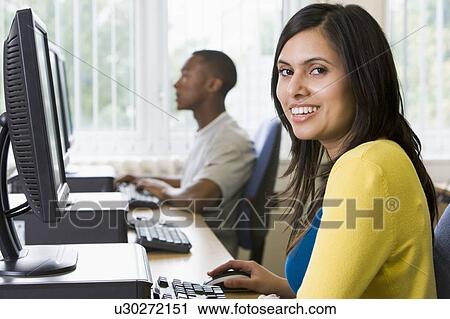Stock Image - Woman sitting at a computer terminal with man in background (selective focus/high key). Fotosearch
