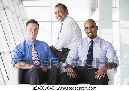 Stock Image - Three businessmen sitting indoors smiling (high key/selective focus). Fotosearch