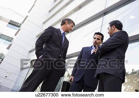 Stock Image - Three businessmen standing outdoors by building talking (high key/selective focus). Fotosearch