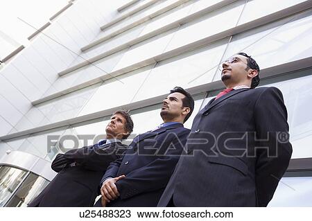 Stock Image - Three businessmen standing outdoors by building (high key/selective focus). Fotosearch