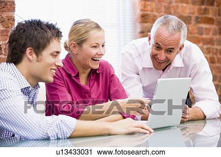 Stock Image - Three businesspeople in office with laptop pointing and laughing. Fotosearch
