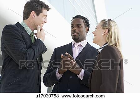 Three businesspeople standing outdoors by building talking and smiling View Large Photo Image Stock Photo - Three businesspeople standing outdoors by building talking and smiling. Fotosearch