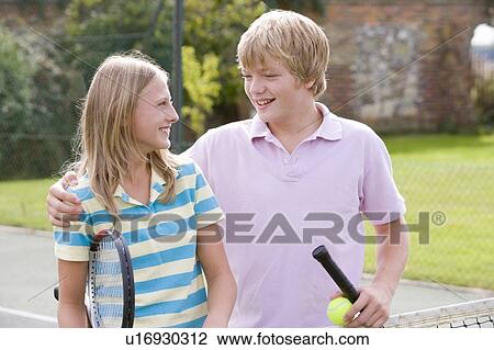 Stock Image - Young couple with rackets on tennis court smiling. Fotosearch