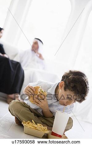 Stock Photo - Young boy with fast food in living room with parents in background (high key/selective focus). Fotosearch